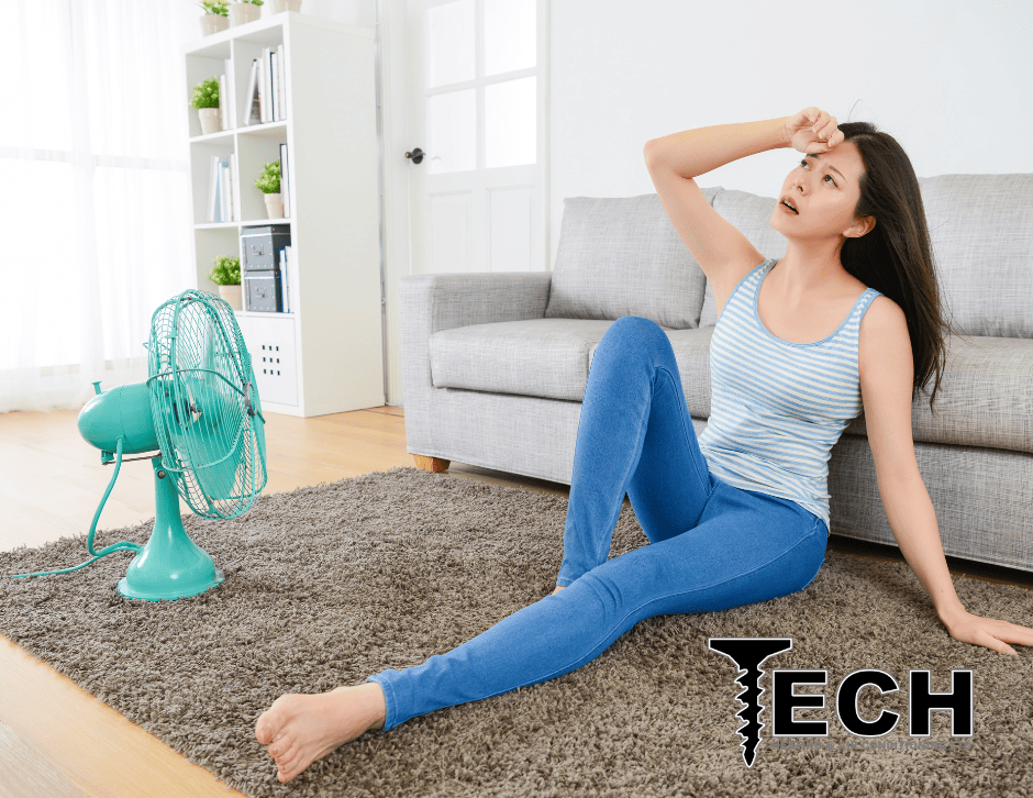 a woman sitting in a hot livingroom with a fan. Is one room in your home hotter than the others? Tech Heating & Air Conditioning in Red Deer can help.